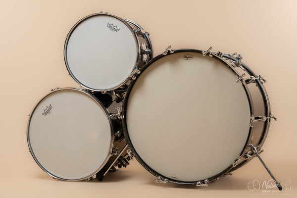 A three-piece drum kit with white drumheads and silver hardware, photographed against a solid beige background. 